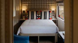 View through a Suite Cabin door, over two blue chairs to with a plush white bed with black and white textile cushions.