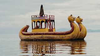 A figure stands on top of an open cabin on a traditional boat with two animal heads made with totora reeds, in still waters.