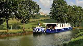 View from a riverbank of the blue and white Fleur de Lys, flying the tricolour flag as it cruises past Château de Châteauneuf.