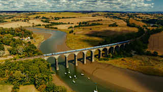An aerial view of a train crossing a stone railway viaduct over a river with small boats moored in the water, surrounded by rolling hills and fields.