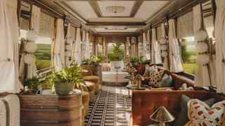 A luxurious vintage train carriage interior with large windows, cream curtains, patterned flooring, plush sofas, plants, and decorative cushions, creating an elegant and cosy atmosphere while the scenery blurs outside.