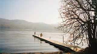 A wooden jetty extends into a calm lake, with two people standing near the end. Ripples spread across the water, and tree branches frame the right side. Distant hills are visible under a hazy sky.