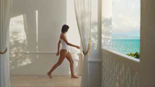 A woman in a white bathing suit walks across the cool spa floor to the terrace where the azure sea is glimpsed over a wall.