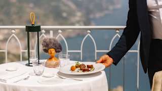 A waitress sets a plate of food down on a terrace table, with a Sandulli figurine, overlooking the sublime Amalfi coastline.