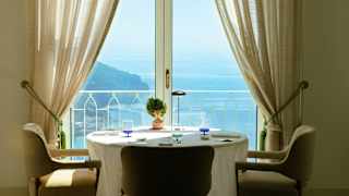 A round table with three chairs sits by glazed doors with tied-back drapes and views out on to the sunny Amalfi coast.
