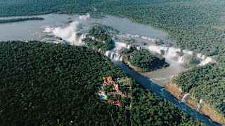 Aerial view of a lush green forest with a wide river and large waterfalls, with a cluster of buildings near the water’s edge. Mist rises from the falling water, blending into the dense forest landscape.