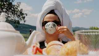 A woman with a towel hair-wrap drinks tea from a porcelain botanical-print cup, seen at eye-level over a table of pastries.