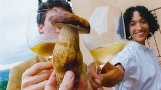 A hand holds up a big porcini mushroom as a man in yellow and a woman in white toast it with glasses of chilled white wine.