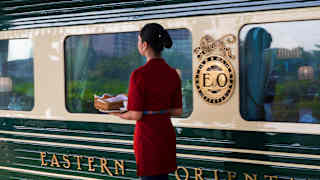 A female staff member in a red top and blue skirt carries a tray along a platform alongside the Eastern and Oriental Express.