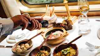 A waiter's hands crack a black pepper mill over four small dishes of brunch food, seen at a window table with a white cloth.