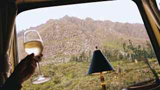 A hand holds a glass of white wine inside a train, with a mountain landscape visible through the window and a small lamp on the table.