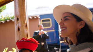 A woman in a sun hat and patterned poncho stands next to an Indigenous woman in traditional clothing and a red hat, with a railway carriage and a man in a suit in the background.