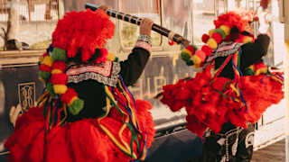 Two dancers in black trousers and flared red fluffy skirts, hats and pompoms, and holding flutes perform on a train platform.