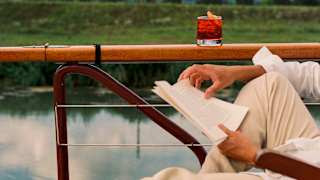 A person in light clothing reads a book whilst reclining on a deckchair by a lake; a red drink rests on the wooden railing, with trees and a cloudy sky in the background.