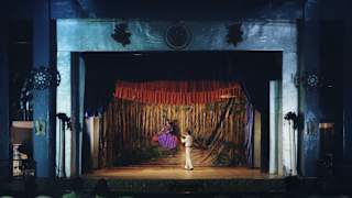 A magician in white levitates a woman in a purple on a red cushion on stage during The Great Mandrake show, seen from afar.