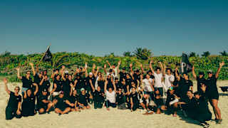 A group of people pose at the back of a sunny beach in black and white casual wear, waving their arms and two Parley flags.
