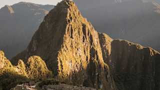 Green-gold with sun, Huayna Picchu looms over the shaded Machu Picchu ruins in a breathtaking aerial view of the citadel.