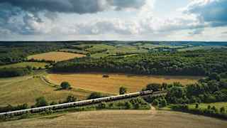 Gently rolling hills reveal a patchwork of yellow and green fields and woods. In the foreground the elegant steam train snakes