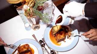A waiter's gloved hand pours gravy over crisp Beef Wellington as a couple indulge in seasonal fine dining, seen from above.