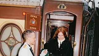 A uniformed train steward holds the hand of a red-haired woman in a black coat as she steps down from the Zena carriage.