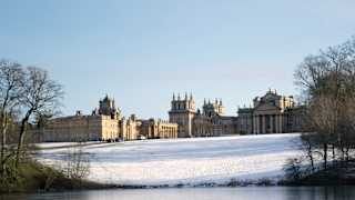 Blenheim Palace, in winter sun, sits atop a gentle sloping field covered with snow, which reflects on the icy lake water below.