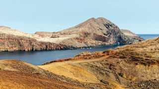 View over a rocky ridge with gold grasses to a deep blue inlet where four small boats are anchored by Ponta de São Lourenço.