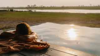 Eye-level view of a man in the pool with his head resting on his arms on the edge, with a towel and glass of water.