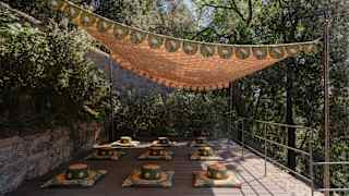 A ethnic brown and green sail shades nine floor cushions and pillows spaced evenly on a tiled terrace surrounded by trees.