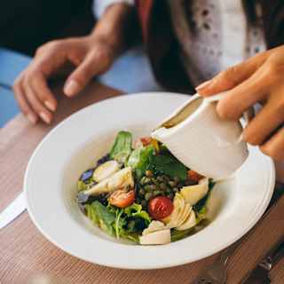 A woman pours a jug of dressing over her vibrant salad with leaves, tomato, artichoke hearts and avocado, seen from above.
