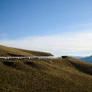 The Andean Explorer train travels across a grass hill in a distant view capturing a lake with a mountainous border.