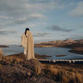 A woman wearing a white woollen shawl watches the train from a high rock as it passes Lake Saracocha below, seen from behind.