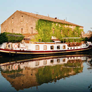 The Alouette, reflected in the still Canal du Midi waters, waits outside the boating station by a rustic, ivy-clad building at Le Somail.