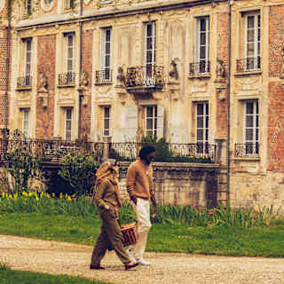 A woman and man, wearing ochres and browns, stroll along a path by Château de Longecourt-en-Plaine, carrying a picnic hamper.