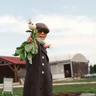 Chef Dominique Crenn, in dark glasses and a flat cap, stands in a vegetable farm, holding up a bunch of fresh-pulled turnips.
