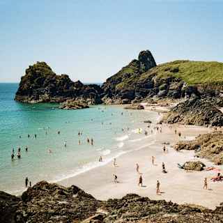 People relax and swim at a sandy beach surrounded by rocky cliffs and clear blue water under a sunny sky. Green hills rise in the background.