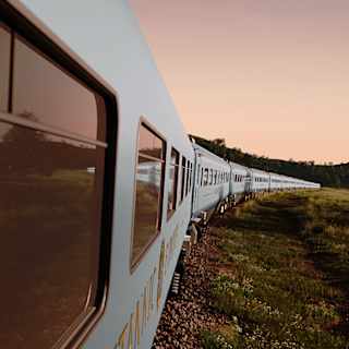 A long passenger train curves through a grassy landscape at sunset, with wildflowers and small rocks along the railway. The sky is soft orange and pink, and the scene feels calm and peaceful.