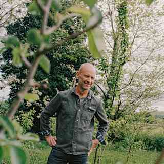 A smiling bald man in a denim jacket stands outdoors in a grassy area with trees and greenery in the background, partly framed by tree branches in the foreground.