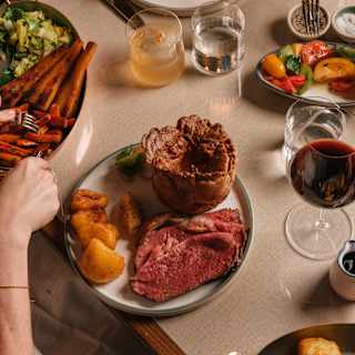 A guest reaches for carrots to add to a sumptuous plate of roast beef, potatoes and Yorkshire pudding, served with sides and wine.