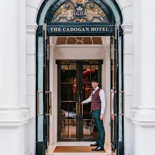 A male staff member with a maroon waistcoat and bow-tie motions towards the double doors in The Cadogan's porch entrance.