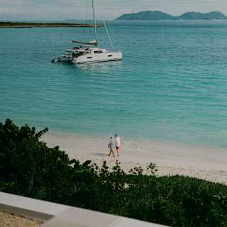 A luxury catamaran floats in the azure bay waters, seen from a balcony over the beach, where two figures walk along the sand.