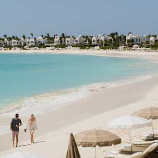 High angle view over the curve and bright blue water of Maundays Bay, where two guests stroll along the soft cream sand.