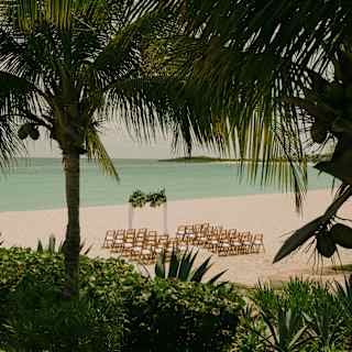 View from above through palm trees to a beach where two columns of chairs face a wedding arch with a sublime sea backdrop.