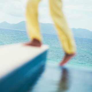 A guest in yellow trousers, seen waist-down in soft-focus, climbs up onto a pool edge in the foreground, with a sea backdrop.