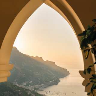 An archway frames a view of the silvery Amalfi coastline beneath an apricot sky as the sun fizzles behind a mountain ridge.