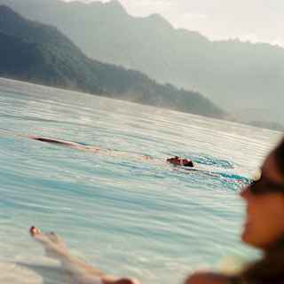 A man swims on his back in the pool, seen over the shoulder of a woman in soft-focus in an image that captures the hill views.