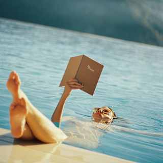 A woman floats on her back, reading a book in the pool with her feet up on the pool's edge, seen close-up from the poolside.