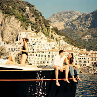 A man an woman sit with legs over the prow of a boat while two others bathe on deck, as they enjoy a cruise past Amalfi town.