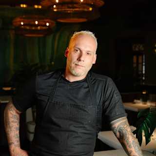 Restaurant Y Chef Luiz Filipe Souza, blond with tattoos, wears a black shirt and apron and leans on a table with one arm.