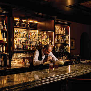 Sub-lit bottle shelves gleam behind a barman as he concentrates on preparing a cocktail at Bar Visconti, taken in low light.
