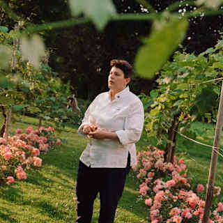 Head Chef Vania Ghedini, in black trousers and white shirt, stands on a garden pathway lined with vines and pink roses.